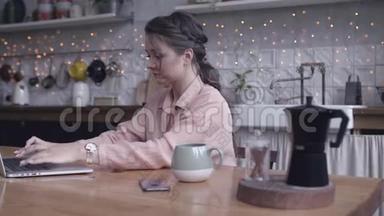 集中精力的年轻女子坐在厨房的木桌旁，在她的<strong>银色笔记本</strong>电脑上用茶或